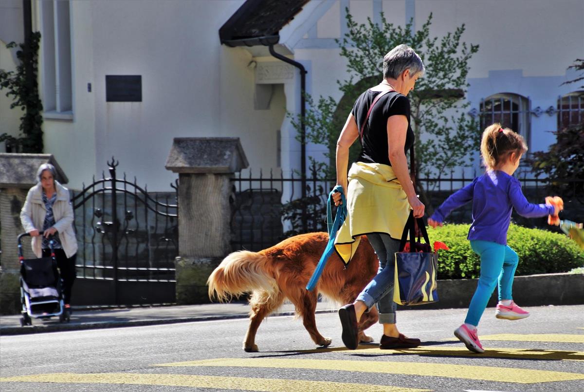 Kvinne med hund går over gata saman med eit barn. I bakgrunnen eldre person med rullator. - Klikk for stort bilete